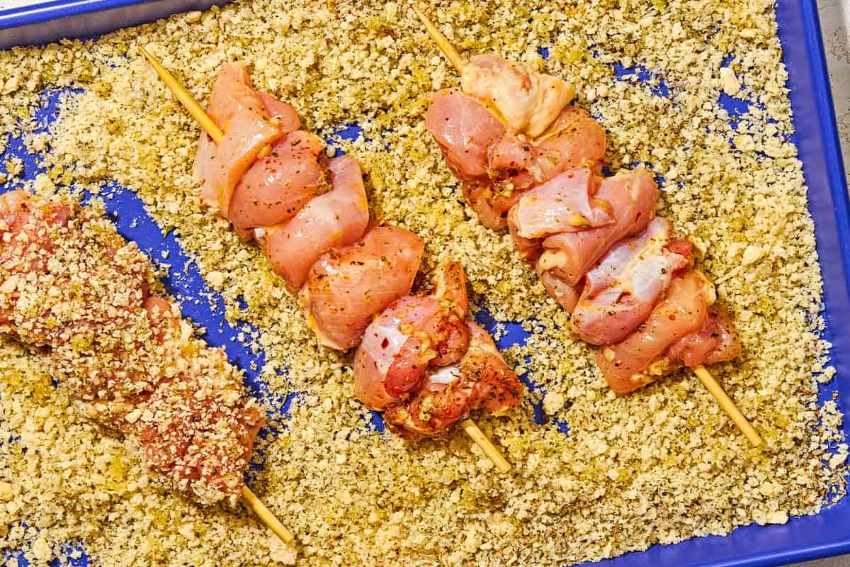 3 uncooked chicken spiedini skewers being coated with breadcrumbs on a sheet pan.