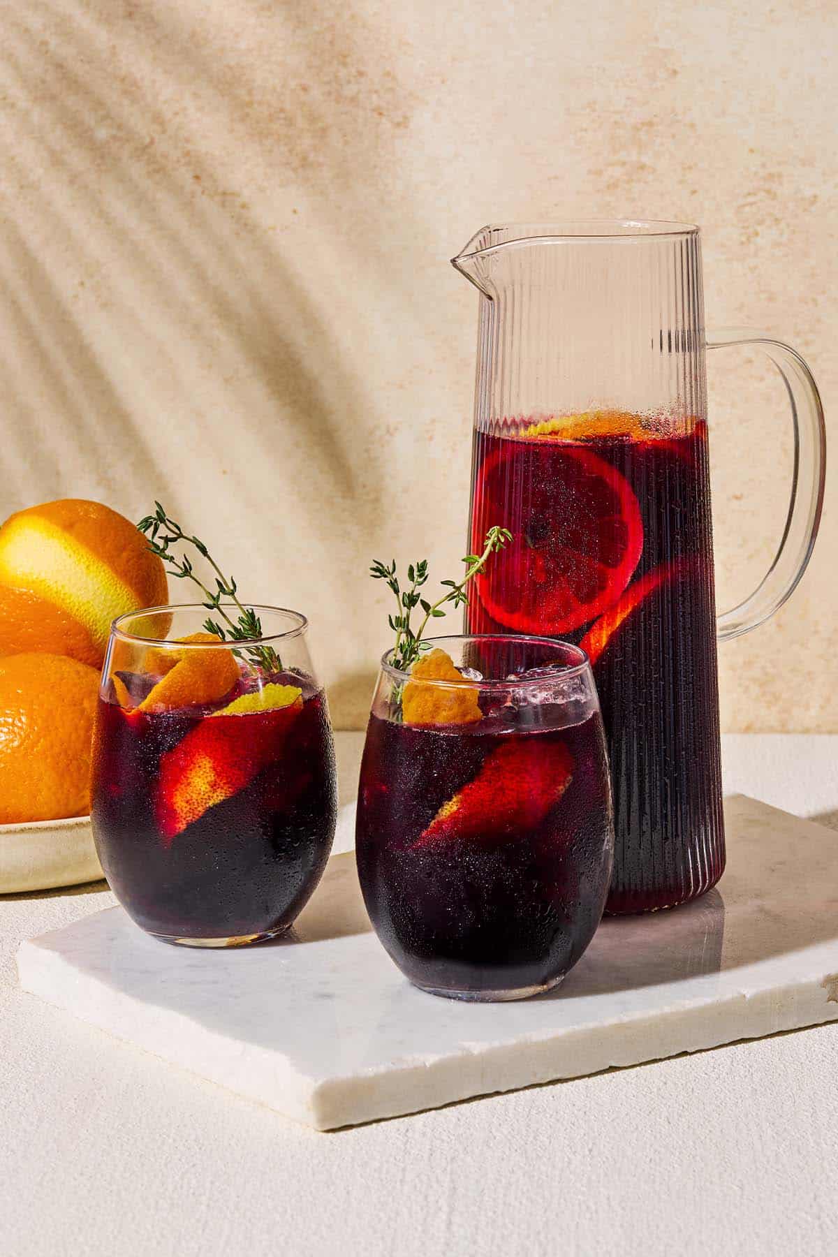 A pitcher non alcoholic sangria next two glasses of the sangria garnished with sprigs of fresh thyme and orange and lemon peels on a marble serving tray. Behind this is a bowl of oranges.